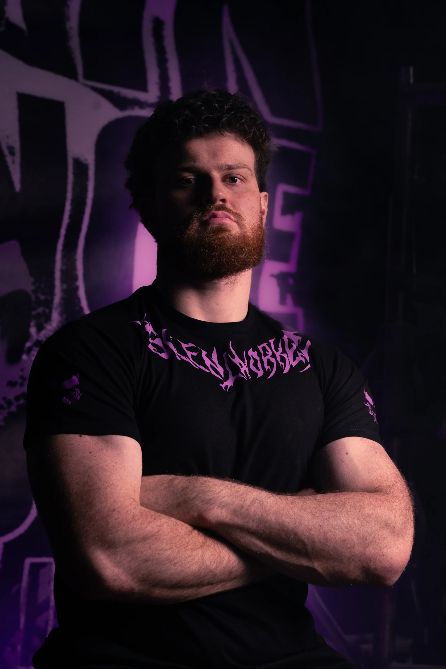 Muscular man in Silent Worker powerlifting apparel with arms crossed, purple gym backdrop
