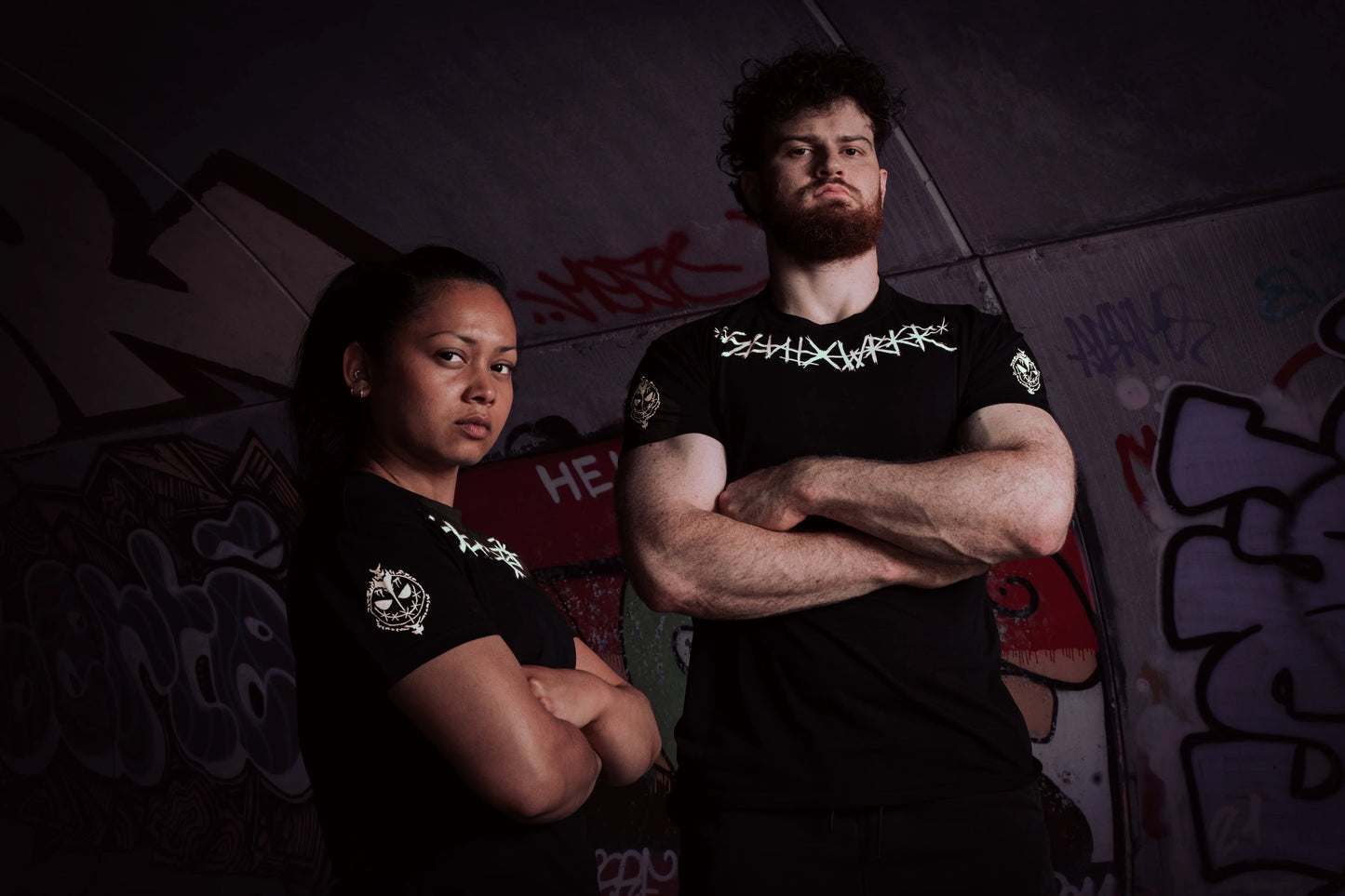 Two athletes in Silent Worker powerlifting apparel posing with arms crossed in a graffiti tunnel